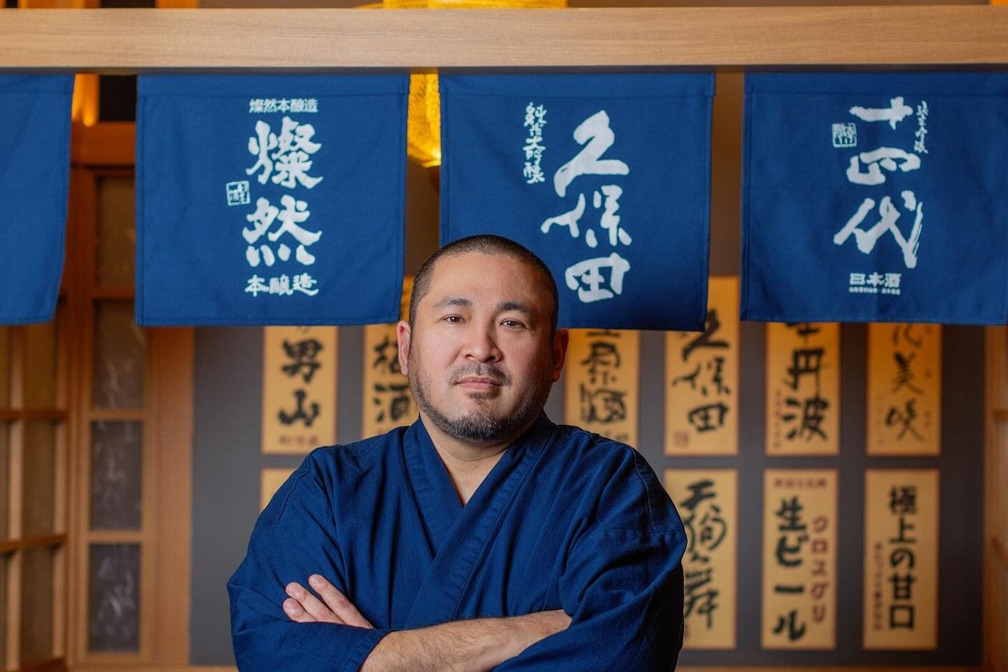 Junzo Miyajima 主厨双臂交叉,站在 Genki Izakaya 的清酒旗幡和手写菜单牌下