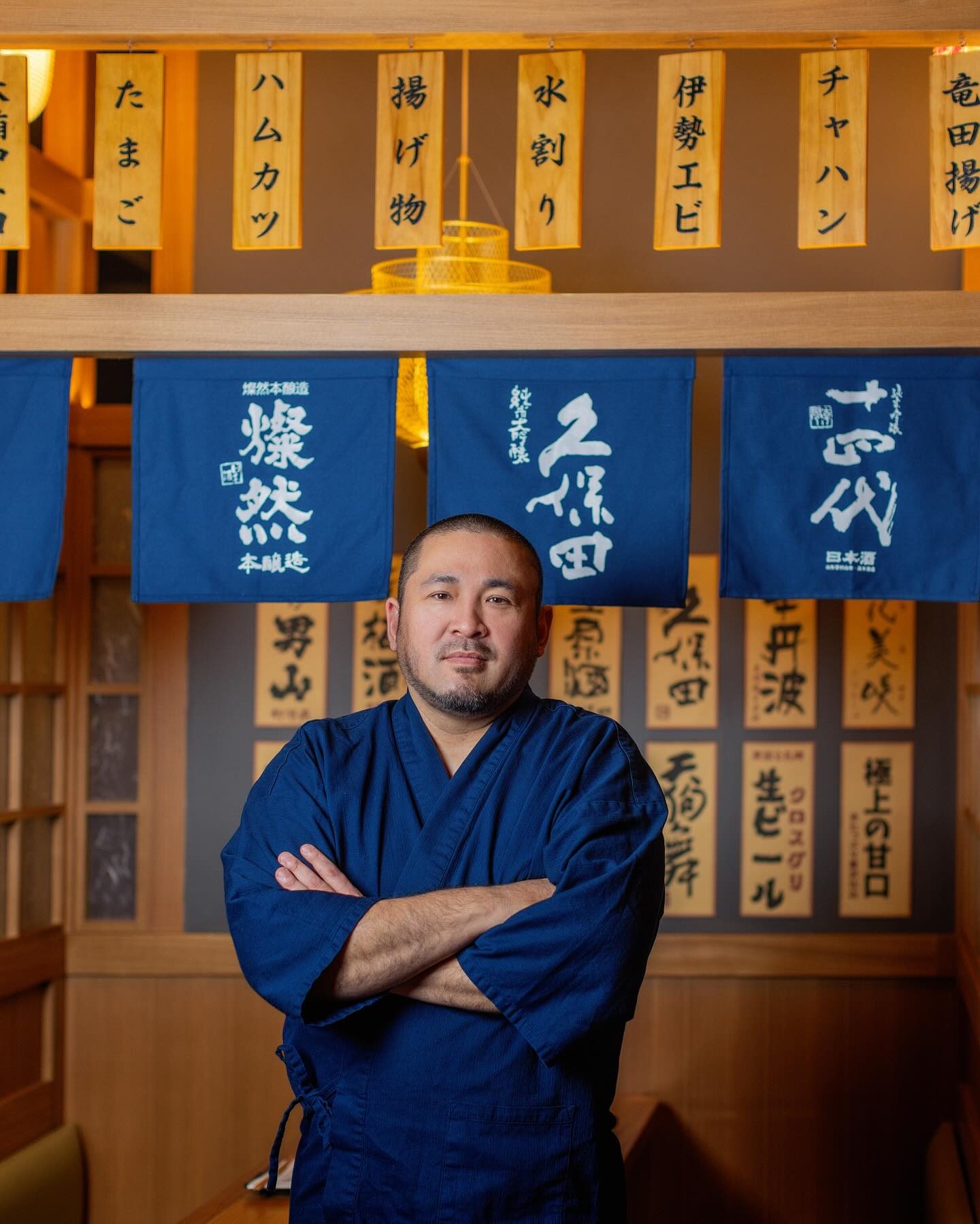 Chef Junzo Miyajima, arms crossed, under sake banners and handwritten menu tags at Genki Izakaya