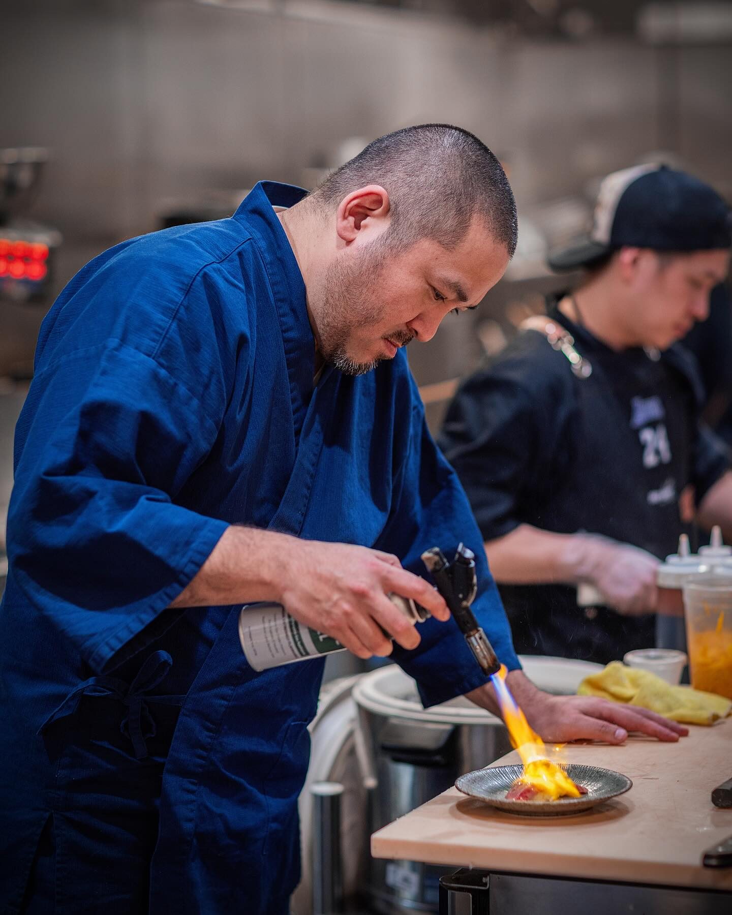 Chef Junzo Miyajima in a blue samue jacket using a kitchen torch to sear a small plate of fish at a wood counter