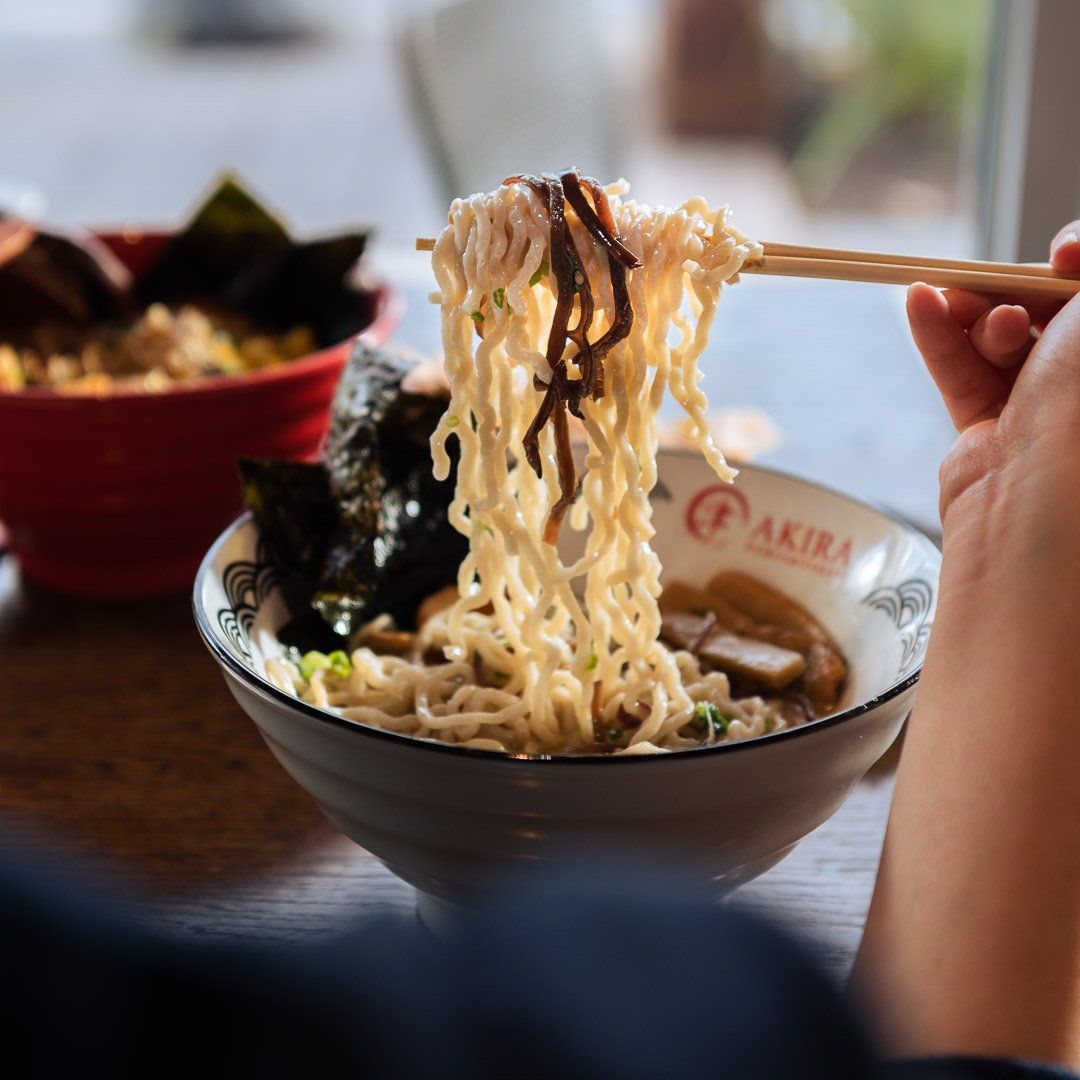 Chopsticks lifting Akira Ramen's curly noodles, made in house daily, from a bowl of ramen